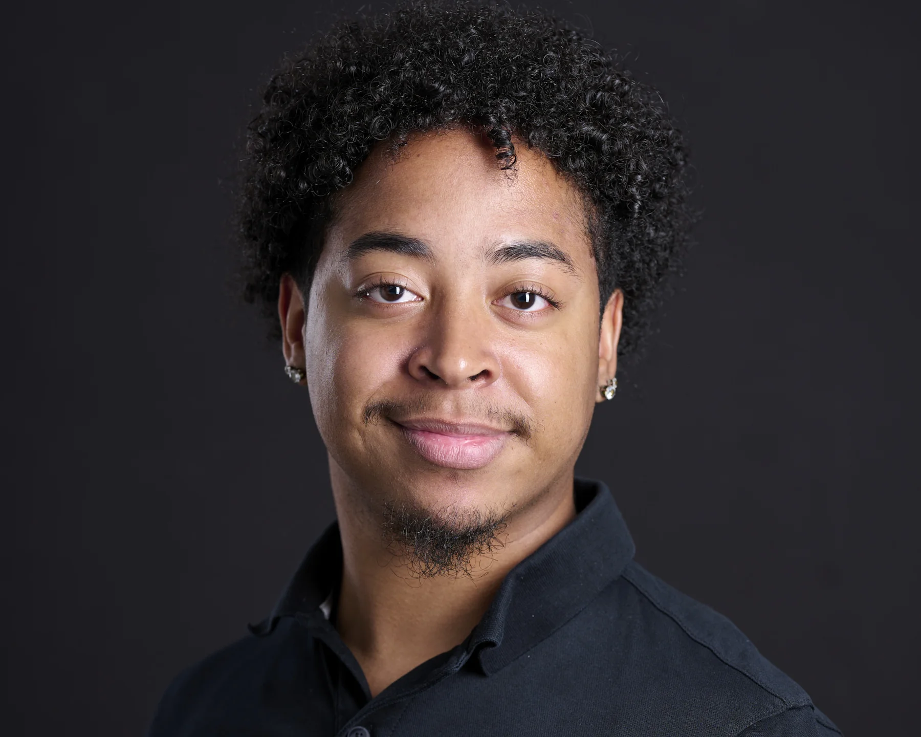 Young professional male actor with curly hair and diamond studs smiling warmly in black polo against dark backdrop
