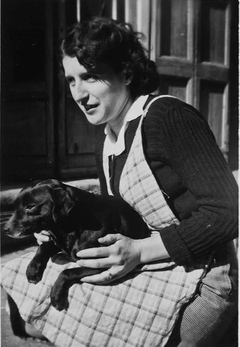 Black and white photograph by Vivian Maier showing Madeleine with natural expression looking to the side, cradling black dog Mirou gently in her arms, wearing dark cardigan with white collar and plaid apron, seated outdoors with wooden building behind her, French Alps 1959