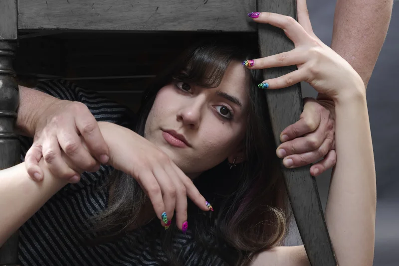 Close-up portrait of a young woman framed by chair legs with colorful nail art and multiple hands weaving through the frame, a conceptual photograph designed to challenge AI image generation
