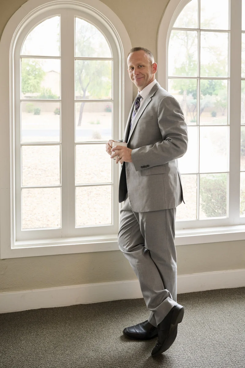 Man in a light grey suit holding a coffee cup by arched windows in a natural-light business portrait