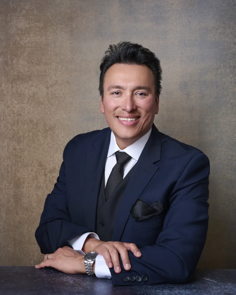 Man in navy three-piece suit with black tie and pocket square leaning forward with hands clasped against textured brown backdrop executive headshot Phoenix Arizona