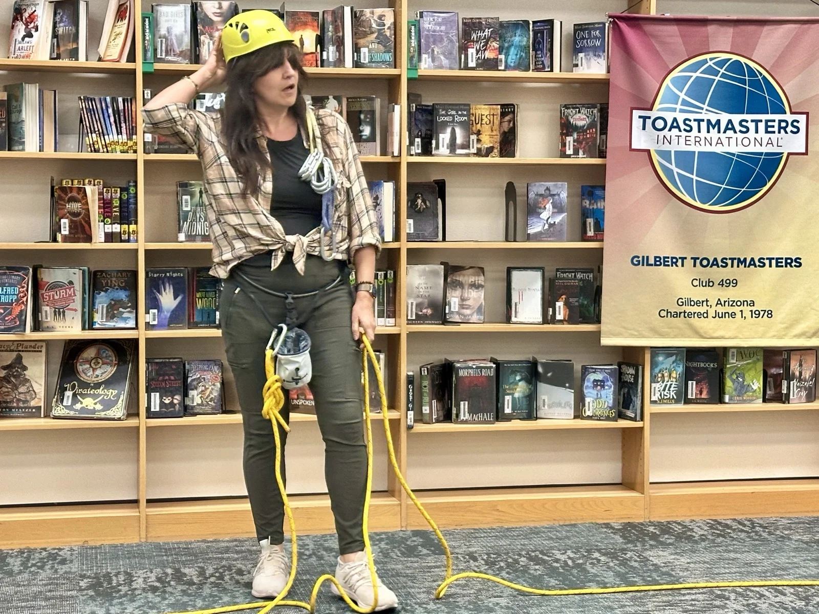 Phoenix headshot photographer Marie Feutrier telling her Tall Tale at Gilbert Toastmasters in a yellow climbing helmet, with a climbing rope at her feet and the club banner beside her