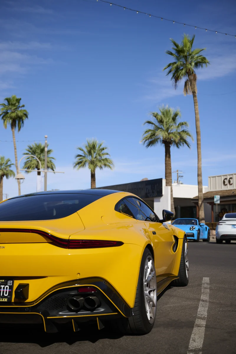 Yellow Aston Martin Vantage parked on a Scottsdale street with palm trees and a blue Porsche in the background