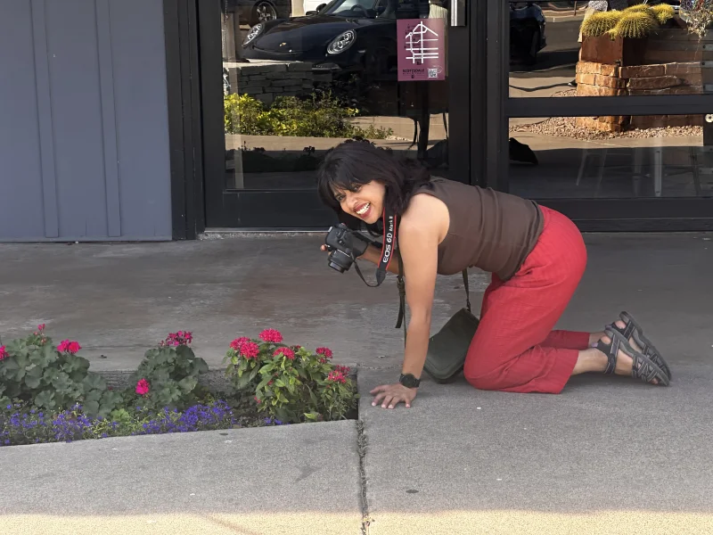 Photographer Soumya kneeling on a Scottsdale sidewalk with her Canon camera, photographing a flower bed of pink and red geraniums