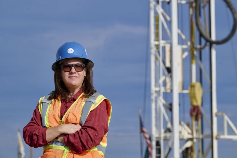 Hydrogeologist in a blue hard hat and orange safety vest standing in front of a drill rig for a personal branding portrait