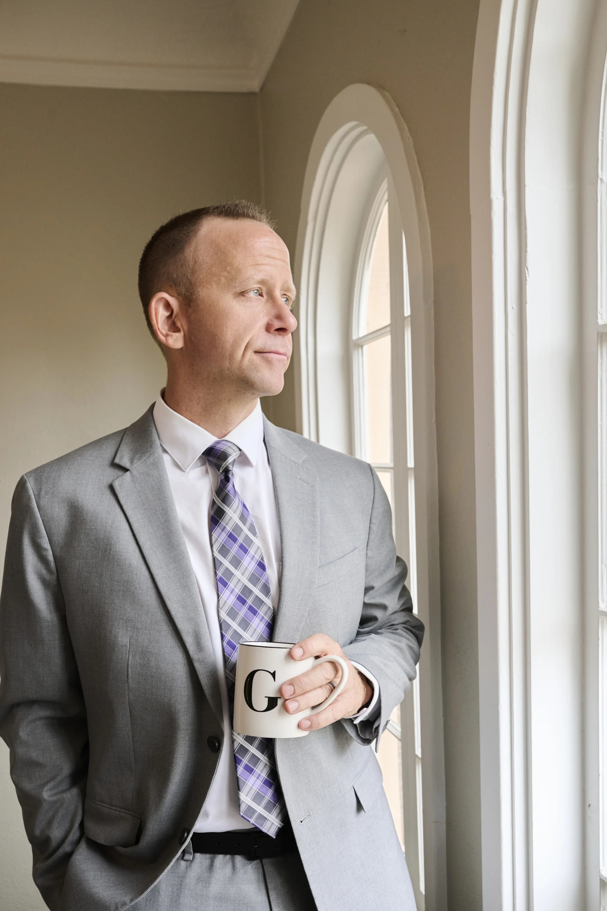Professional male attorney in gray suit holding coffee mug by arched window with natural light