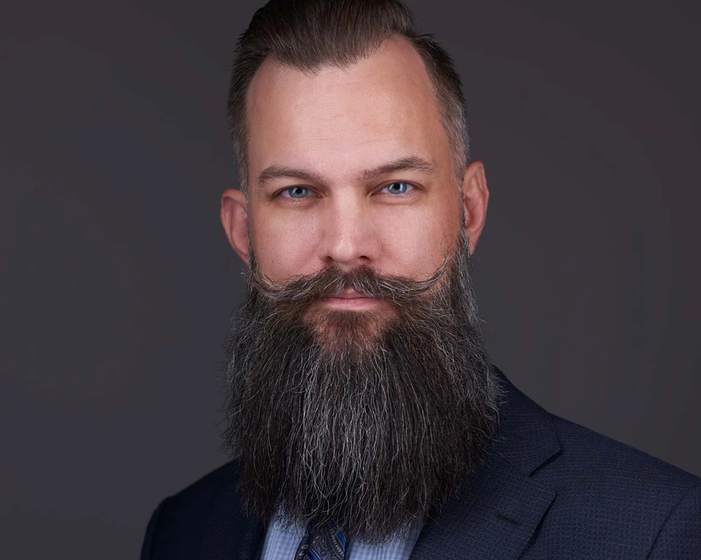 Man with well-groomed beard and styled hair in navy suit professional headshot against grey backdrop