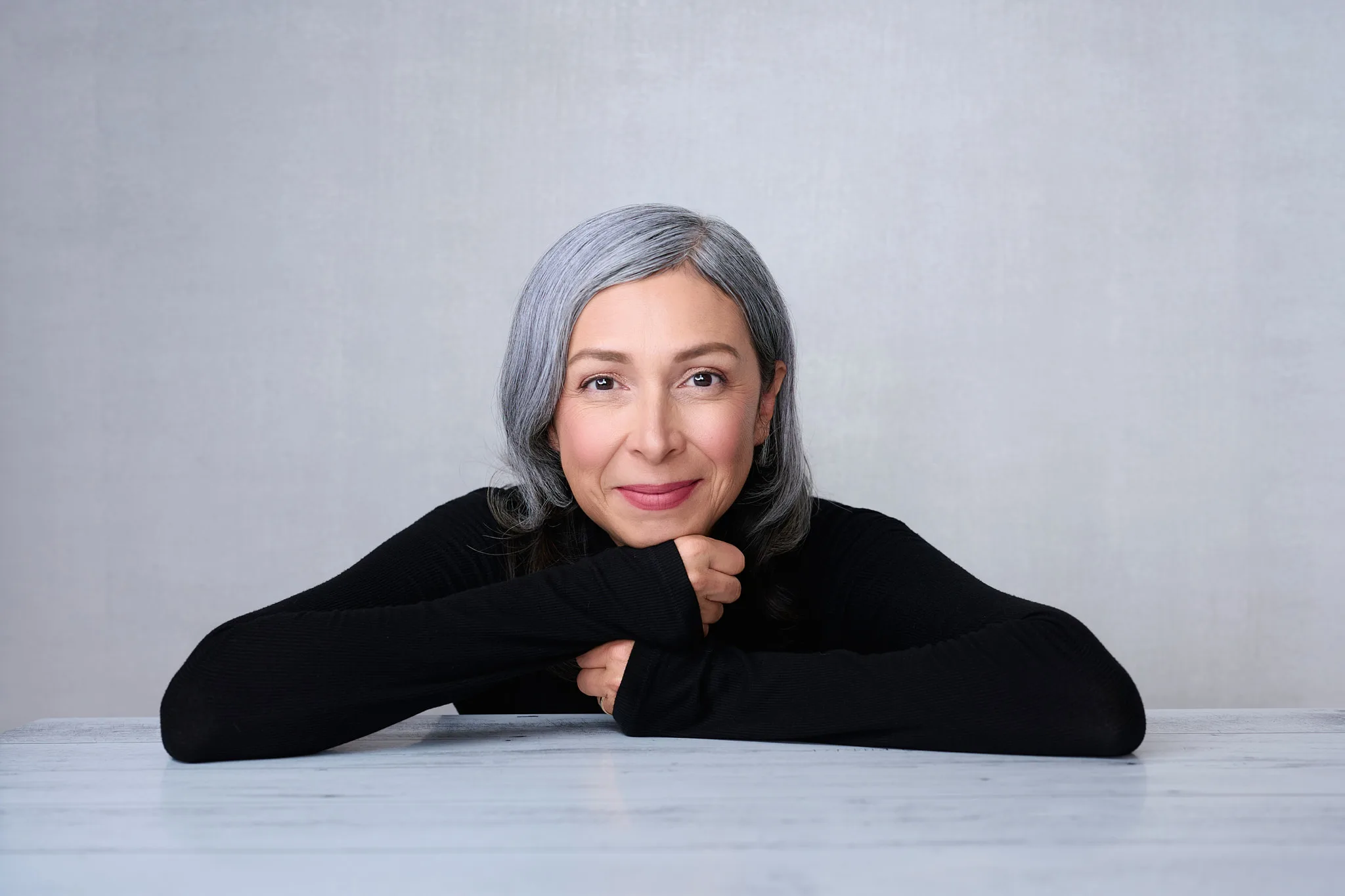 Professional woman with silver hair in black sweater smiling warmly with chin resting on hands