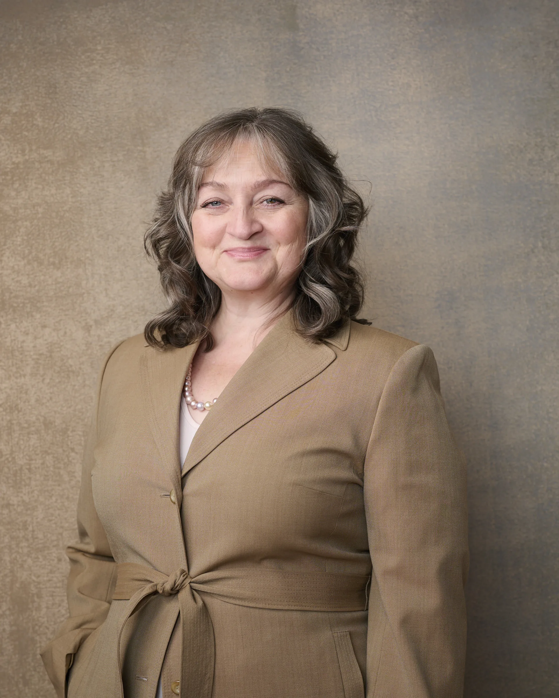 Professional woman in tan blazer with pearl necklace smiling warmly against textured beige backdrop