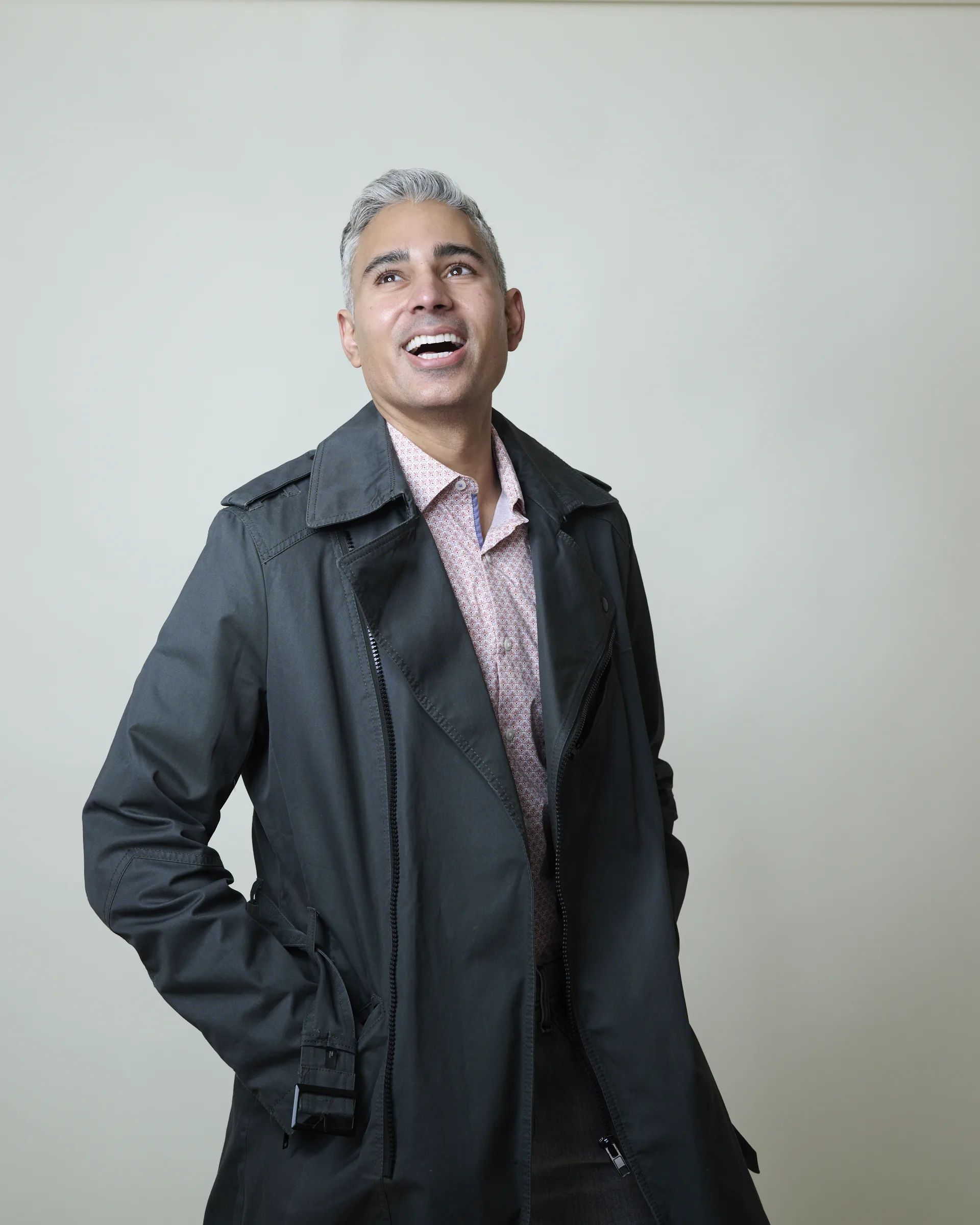 Man with silver hair laughing naturally in dark trench coat and patterned shirt during relaxed portrait session