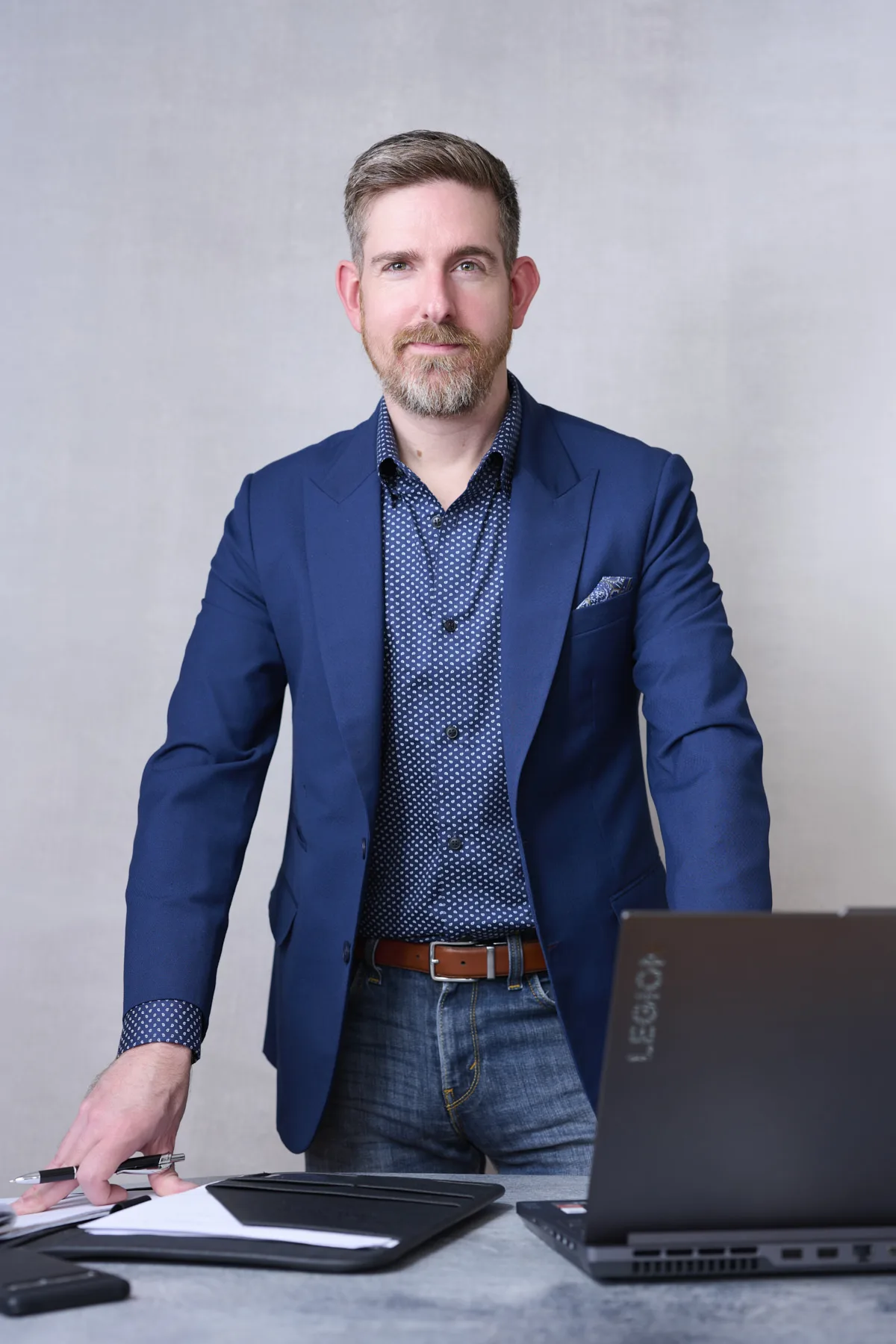 Professional man in navy blazer and patterned shirt leaning confidently at desk during business headshot session